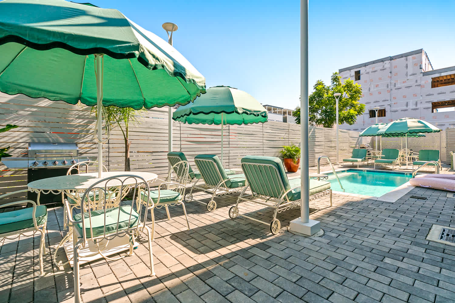 Sun-soaked urban poolside patio with teal-green umbrellas, cushioned lounge chairs and a dining set on a paver deck beside a small rectangular swimming pool and stainless grill.