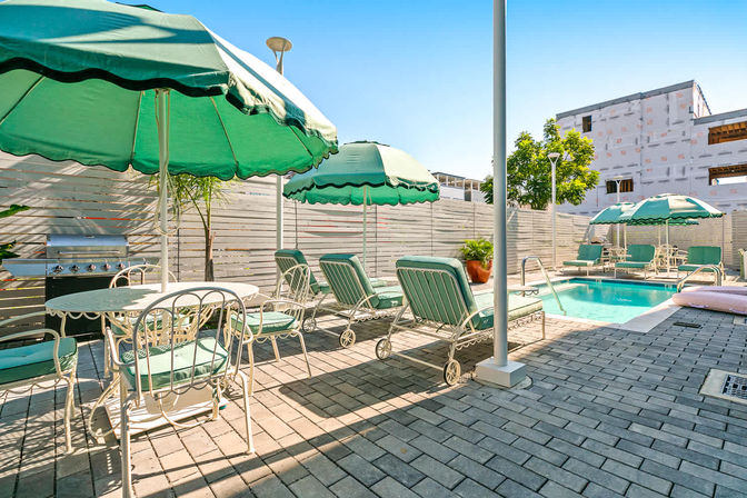 Sun-soaked urban poolside patio with teal-green umbrellas, cushioned lounge chairs and a dining set on a paver deck beside a small rectangular swimming pool and stainless grill.