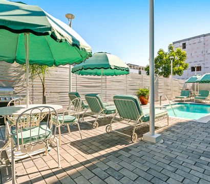Sun-soaked urban poolside patio with teal-green umbrellas, cushioned lounge chairs and a dining set on a paver deck beside a small rectangular swimming pool and stainless grill.