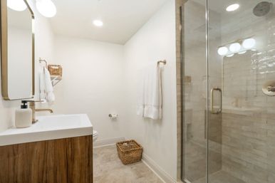 Bright modern home bathroom with glass-door tiled shower, wood vanity and white sink, brass fixtures, white towels and woven storage baskets — crisp, neutral design