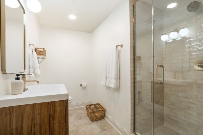 Bright modern home bathroom with glass-door tiled shower, wood vanity and white sink, brass fixtures, white towels and woven storage baskets — crisp, neutral design