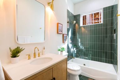 Sunlit contemporary bathroom with white quartz vanity, brass faucet and sconce, rounded mirror, green vertical subway-tiled tub-shower, small potted plants, and clerestory window