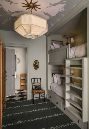 Cozy narrow bedroom with built-in sage-green bunk beds and ladder shelving, vintage chair, pleated octagonal pendant light, decorative ceiling wallpaper, and a doorway revealing a black-and-white checkerboard bathroom floor.