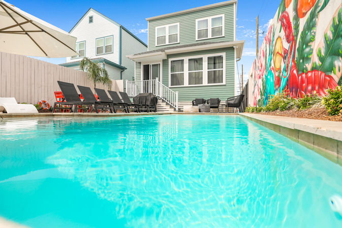 Sunny backyard turquoise swimming pool with black lounge chairs and umbrella, steps up to a two-story mint-green house and a vibrant mural along the fence.