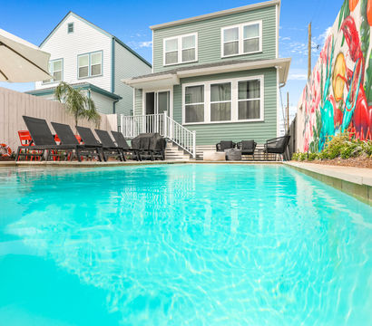 Sunny backyard turquoise swimming pool with black lounge chairs and umbrella, steps up to a two-story mint-green house and a vibrant mural along the fence.