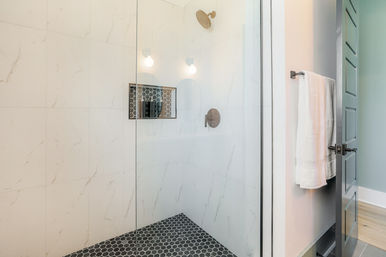 Sleek glass walk-in shower with white marble-look wall tile, black hexagon floor tile, brushed brass rain showerhead, mosaic-tiled recessed niche with bottles, and a white towel on the open gray bathroom door.