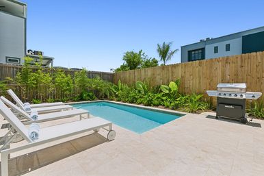 Sunny suburban backyard with a rectangular plunge pool, three white chaise lounges with rolled towels on a tiled patio, lush tropical plants along a wooden fence, and a stainless-steel gas grill ready for poolside entertaining.