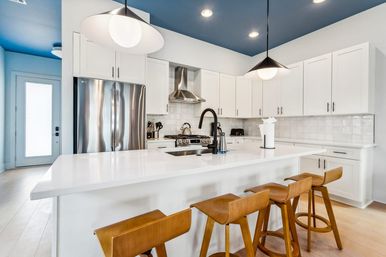 Bright, modern open-concept kitchen with white cabinets and quartz island, stainless-steel fridge and range, navy-blue ceiling, black gooseneck faucet, pendant lights and four wooden bar stools.