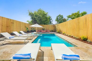 Sunlit private backyard pool with turquoise water, stone deck, white lounge chairs with blue-striped towels, umbrella and a pink outdoor sofa framed by a tall wooden privacy fence.