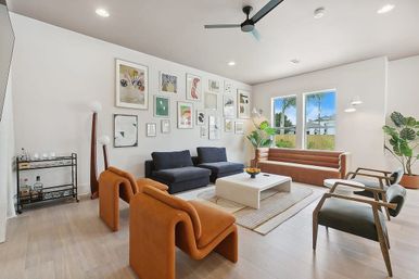 Bright modern living room with gallery wall, navy sectional, tan leather sofa, sculptural orange lounge chairs, mid-century wood armchairs, white coffee table on rug, ceiling fan, indoor plants and large windows overlooking a sunny backyard.