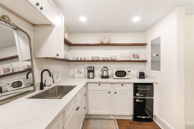 Bright modern white kitchenette with marble-look counters, stainless sink and black faucet, wooden floating shelves stocked with dishes, a coffee station with kettle and microwave, and a small black retro mini refrigerator.