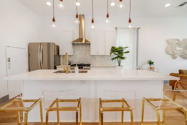Bright modern white kitchen with large island and brass faucet, hex-tile backsplash, stainless refrigerator and range, exposed-bulb pendant lights, clear acrylic bar stools with gold frames, and a potted plant.
