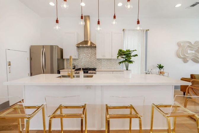 Bright modern white kitchen with large island and brass faucet, hex-tile backsplash, stainless refrigerator and range, exposed-bulb pendant lights, clear acrylic bar stools with gold frames, and a potted plant.