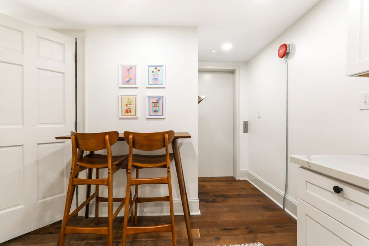 Cozy modern apartment nook with two mid-century wooden bar stools at a narrow breakfast bar, four colorful framed prints on a white wall, warm hardwood floors and white kitchenette cabinetry.
