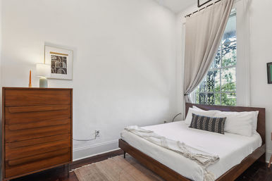 Bright, minimalist bedroom in a city apartment: mid-century wooden bed with white linens and textured throw, matching tall dresser with lamp and candle, large window with light gray curtain and decorative wrought-iron balcony, hardwood floors and abstract wall art.