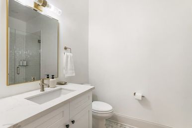 Bright minimalist white bathroom with marble-look vanity countertop, undermount sink, brushed-nickel faucet, gold-framed mirror reflecting a glass-enclosed subway-tile shower, wall towel, toilet, and patterned floor tile.