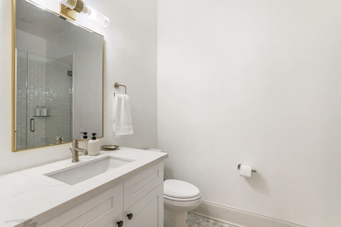 Bright minimalist white bathroom with marble-look vanity countertop, undermount sink, brushed-nickel faucet, gold-framed mirror reflecting a glass-enclosed subway-tile shower, wall towel, toilet, and patterned floor tile.