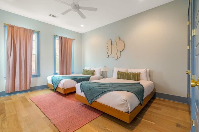Sunlit twin guest bedroom with two low wooden platform beds dressed in white linens, teal throws and green lumbar pillows, blush curtains over tall windows, pink area rug on hardwood floors, ceiling fan and abstract wall art.