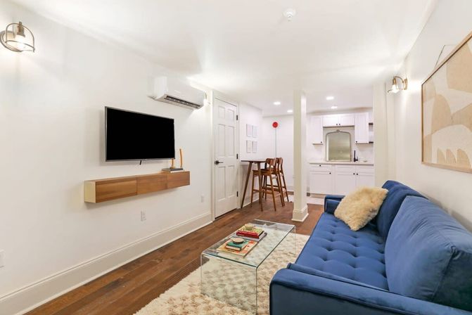 Cozy modern basement living space with a navy tufted sofa and fuzzy pillow, wall-mounted TV above a floating wood console, glass coffee table on a shag rug, hardwood floors, and a compact white kitchenette with bar stools.