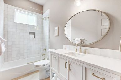 Bright spa-like modern bathroom with white tiled tub-shower and frosted window, white vanity with sink, brushed-nickel faucet and large oval mirror.