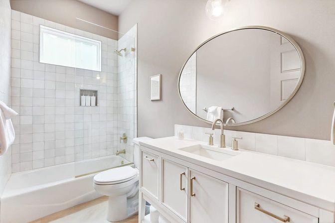 Bright spa-like modern bathroom with white tiled tub-shower and frosted window, white vanity with sink, brushed-nickel faucet and large oval mirror.