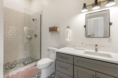 Bright modern home bathroom with frameless glass subway-tile shower, pebble-pattern mosaic floor, white toilet, gray vanity with white quartz countertop and brass hardware, wall mirror and wicker shelf.