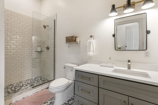 Bright modern home bathroom with frameless glass subway-tile shower, pebble-pattern mosaic floor, white toilet, gray vanity with white quartz countertop and brass hardware, wall mirror and wicker shelf.