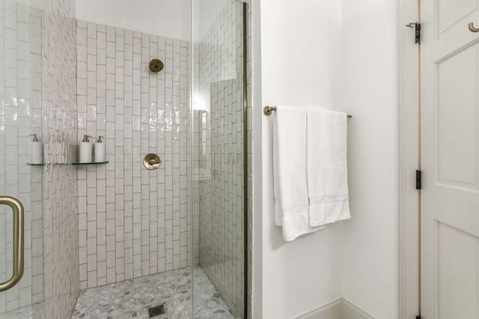 Glass-enclosed modern bathroom shower with vertical white subway tiles, brass showerhead and control, patterned gray hex floor tiles, glass shelf with soap dispensers, and a folded white towel on a wall-mounted bar.