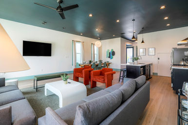 Open-concept modern living room and kitchen with a dark teal ceiling, gray sofas, two vibrant orange armchairs, white sculptural coffee table, wall-mounted TV, dining table by windows, and a black kitchen island with pendant lights over hardwood floors.