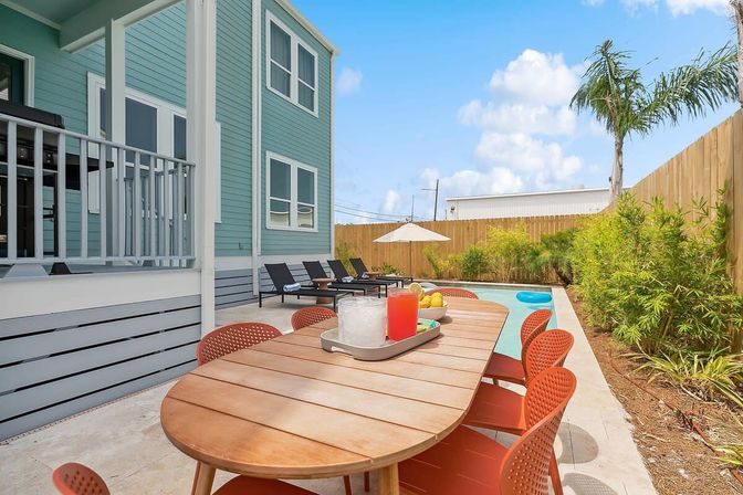 Coastal backyard patio with wooden dining table and red chairs, turquoise two-story house, small rectangular swimming pool with lounge chairs, palm tree and privacy fence under a bright blue sky