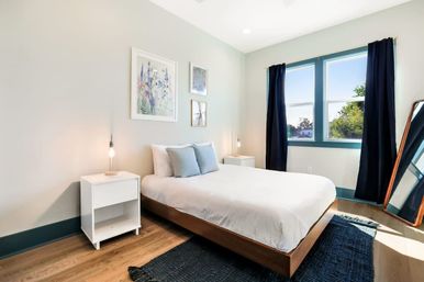 Sunlit modern bedroom with low wood bed, white bedding and blue pillows, white nightstands with hanging bulb lamps, botanical wall art, large window framed by navy curtains, leaning full-length mirror, hardwood floor and dark area rug.