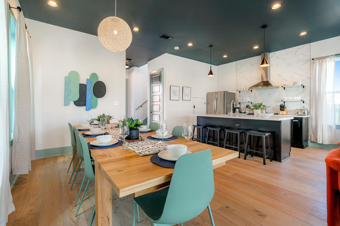 Modern open-plan dining room and kitchen with a wooden dining table set for six, teal chairs, potted centerpieces, black breakfast island with stools, stainless steel appliances, marble backsplash, pendant and recessed lighting, and light hardwood floors.