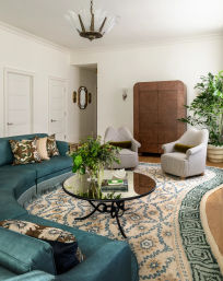 Stylish, bright living room interior featuring a curved teal sectional, round glass-top coffee table on a patterned blue-and-beige rug, two striped armchairs, tall potted plants and a brown armoire.