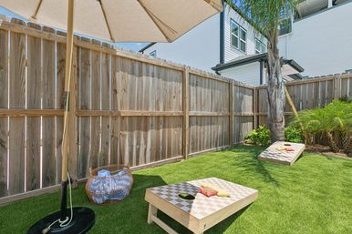 Small fenced backyard with wooden privacy fence, artificial turf, two cornhole boards with colorful bean bags, a patio umbrella, wicker basket and a palm tree beside a modern townhouse