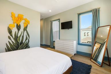 Sunlit modern bedroom with crisp white bed, oversized yellow floral wall mural, blue-trim windows, wall-mounted TV, white dresser, leaning full-length mirror, navy rug, and wood floors