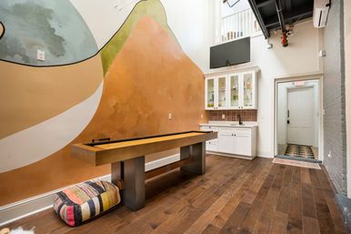 Bright urban loft game room with wooden shuffleboard table, colorful abstract wall mural, hardwood floors, white wet bar with glass cabinets and wall-mounted TV