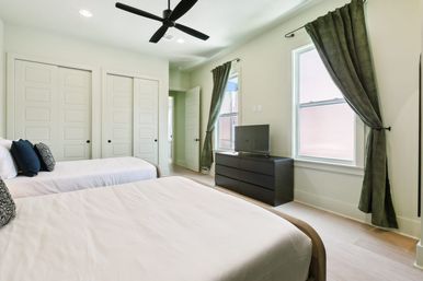 Bright, airy modern twin bedroom with two beds and white bedding, black ceiling fan, hardwood floors, black six-drawer dresser with flat-screen TV, green curtains framing windows, and white paneled closet doors.