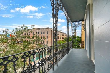 Ornate wrought-iron balcony with decorative posts overlooking a tree-lined urban street and brick commercial buildings under a bright blue sky.