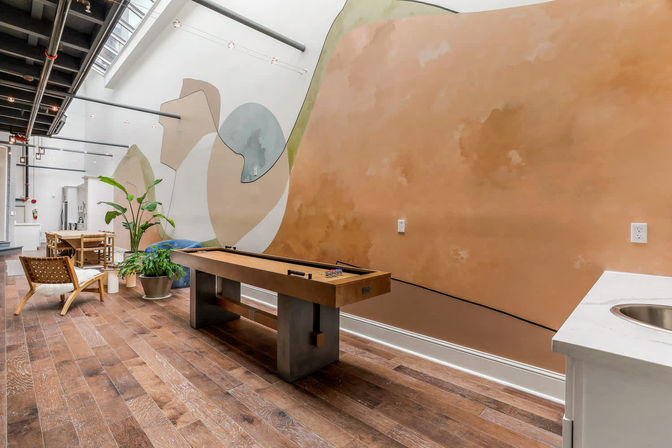 Bright loft-style game room with a wooden shuffleboard table, abstract terracotta-and-pastel mural, wide-plank hardwood floors, skylight and industrial ceiling beams, potted plants and lounge seating.