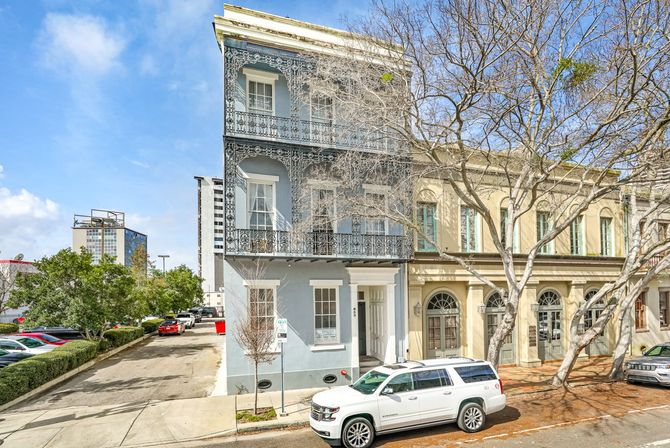 Three-story blue-gray townhouse with ornate wrought-iron balconies in a historic downtown streetscape, leafless trees and a white SUV parked on the sunlit street under a bright blue sky.