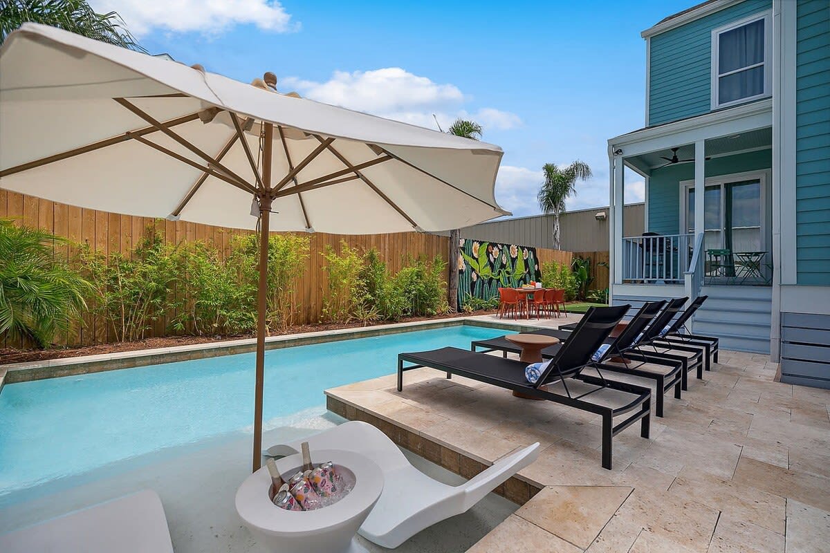 Sunny backyard with rectangular turquoise pool, large white umbrella over a shallow ledge, black chaise lounges on a stone patio, orange outdoor dining set by a wooden fence and a blue two-story house with covered porch.