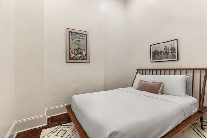 Cozy neutral bedroom with mid-century wooden bed, crisp white linens and dusty-rose lumbar pillow, framed vintage prints on white walls, patterned rug and warm hardwood floor.