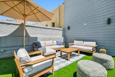 Sunny urban rooftop patio with wooden outdoor sofa set, armchairs and coffee table under a large beige umbrella on green artificial turf, patterned rug and poufs against blue-gray siding.