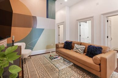 Contemporary living room with tan leather sofa, navy throw pillows, glass coffee table on a patterned rug and a bold geometric accent wall.