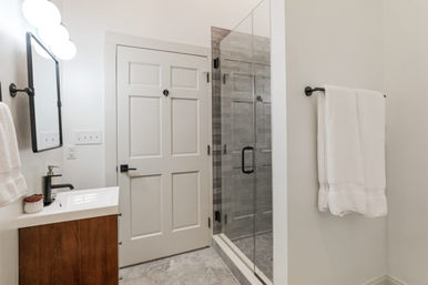 Bright modern bathroom with frameless glass walk-in shower, wood vanity, matte-black fixtures, white towels, and gray tile floor.