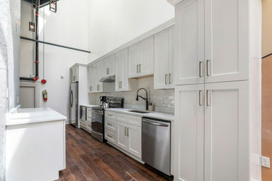 Bright loft-style galley kitchen with white shaker cabinets, quartz countertops, subway tile backsplash, stainless steel appliances, black gooseneck faucet, dark hardwood floors and high ceiling with exposed pipes.