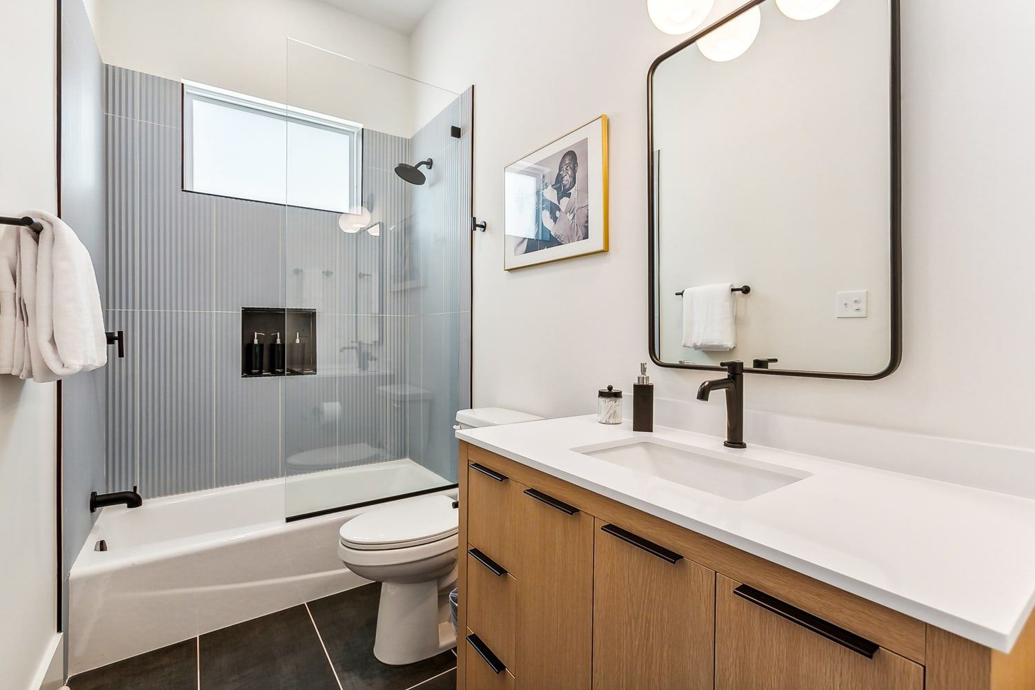 Sleek modern bathroom interior with glass-enclosed tub-shower and ribbed gray tile, white countertop on a wood vanity, black matte fixtures, rounded-rectangle mirror, framed art and folded towels.