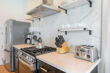 Sleek modern kitchen with stainless steel refrigerator and gas range, marble-look backsplash, kettle on the stovetop, wooden knife block, chrome toaster, and glassware on floating shelves.
