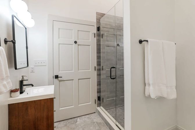 Bright modern bathroom with glass walk-in shower and gray tile, white paneled door, wood vanity with black faucet and mirror, wall-mounted lights and neatly folded white towels on a black towel bar.