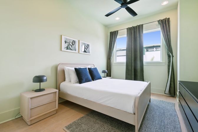 Bright modern bedroom with pale green walls, light-wood bed with white linens and navy pillows, matching nightstand and black lamp, dark curtains over window, ceiling fan and gray rug